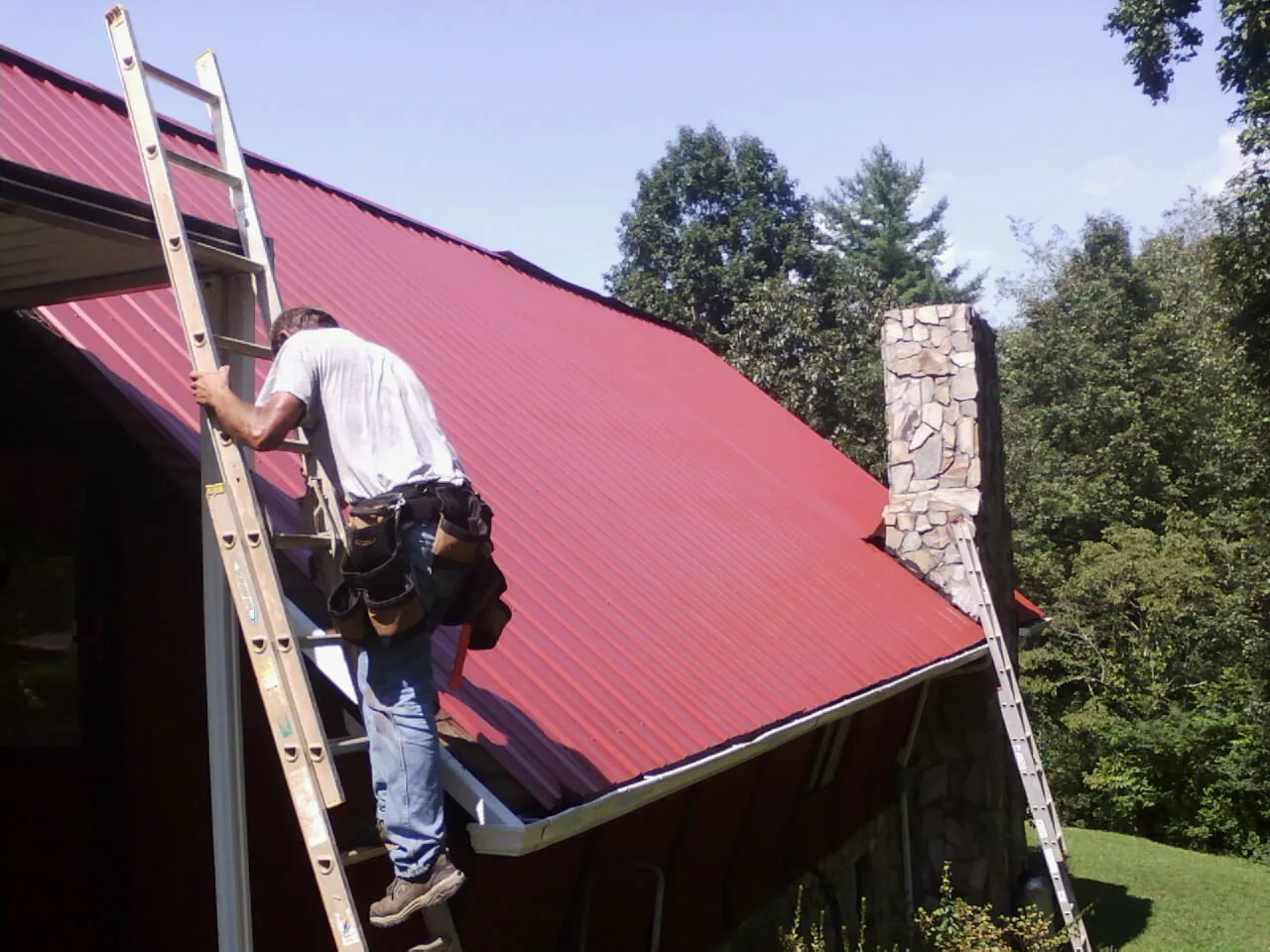 Roofer on a ladder beside a red metal roof and stone chimney — Soffit & Fascia Repair by Roofing Brothers in Bronwood
